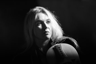 Foto: Susanne Ansorg © S. Ansorg, Foto: Stefan Schweiger Schwarz-Weiß-Aufnahme einer Frau mit offenen, langen blonden Haaren vor schwarzem Hintergrund. An ihrer Schulter lehnt ein Saiteninstrument, dessen hölzerner Steg mit Schnitzereien verziert ist.