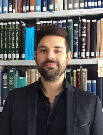 Foto: Andreas Lehnertz © A. Lehnertz Ein Mann mit kurzen dunklen Haaren, dunklen Augen und kurzem dunklen Vollbart steht lächelnd vor einem Bücherregal. Er trägt ein scharzes Hemnd und scharzes Jackett.