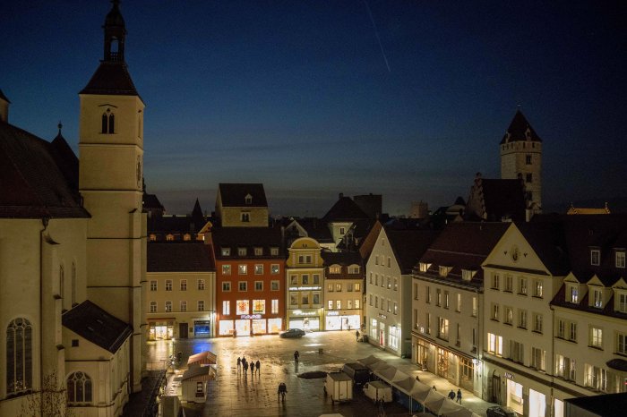 Zu sehen ist ein Platz in der Regensburger Altstadt iin der hereinbrechenden Dunkelheit. Auf der linken Bildseite ist eine Kirche mit heller Fassade zu sehen, das Karree des mit hellem, nass glänzendem Steinpflaster ausgelegten Platzes ist mit ebenfalls überwiegend hell angestrichenen Häusern eingefasst. Auf dem Platz sind einige Zeltbuden aufgebaut, zwischen denen herumlaufende Menschen zu erkennen sind.