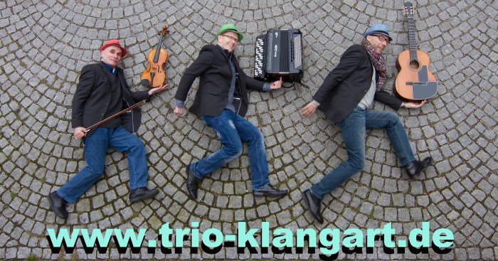 Abgebildet sind drei Musiker des Trios Klangart. Die drei Musiker liegen am Boden und über ihren ausgebreiteten Händen liegen die Instrumente. 