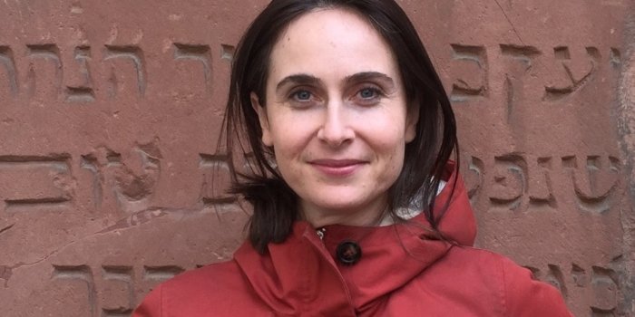 Foto: Naomi Lubrich © N. Lubrich Eine Frau mit dunklen Haaren in rotem Anorak steht vor einer rötlich-braunen Steintafel, auf der hebräische Schriftzeichen eingraviert sind, und blickt in die Kamera.