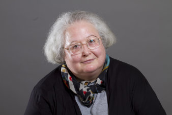 Eine Frau mit grauem, als Pagenkopf geschnittenen volen Haar und einer großen randlosen Brille blickt ernstahft in die Kamera.Über einem hellgrauen Pullover trägt sie eine dunkel Jacke und ein buntes Halstuch.