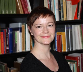 Foto: Annett Martini © A. Martini Eine junge Frau mit kurzen braunen Haaren und braunen Augen blickt lächelnd in die Kamera. Sie trägt einen schwarzen Pullover und sitzt vor einem Bücherregal.