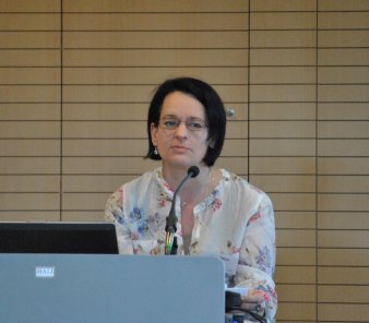 Foto: Birgit Wiedl © B. Wiedl Vor einer hölzernen Paneelwand und hinter einem Pult steht eine Frau und hält einen Vortrag. Sie trägt dunkle halblange Haare, eine schmal rot gefasste Brille und eine helle Bluse mit geblümtem Muster.