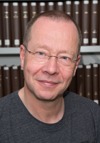 Foto: Christoph Cluse © C. Cluse Ein Mann mit kurzen Haaren, einer randlosen Brille und einem dunkelgrauen T-Shirt blickt freundlich in die Kamera. Im Hintergrund sind Bücherregale zu erkennen.