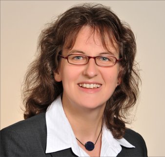 Foto: Claudia Bergmann © C. Bergmann Eine Frau mit mittellangem, gelocktem braunen Haar und einer rot gefassten, rechteckigen Brille lächelt in die Kamera. Sie trägt eine weiße Bluse, ein graues Jackett und eine Kette mit einem blauen kreisförmigen Amulett.
