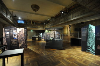 Blick vom Treppenhaus aus in einen Raum mit Holzboden, in dem Ausstellungstafeln und Vitrinen zu sehen sind