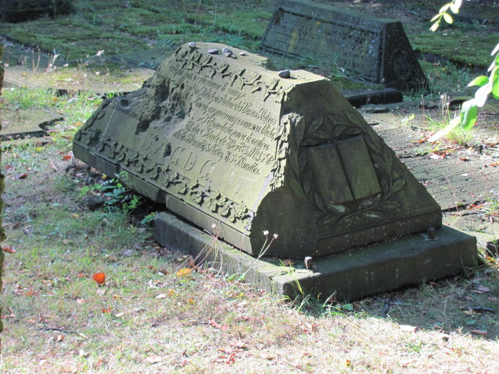 Sephardischer Grabstein auf dem Jüdischen Friedhof Altona Im Sonnenschein ist ein sephardisches Grab zu sehen. Anstelle einer Grabplatte oder eines Grabsteins ist der Sarkophag in Portugiesisch beschrieben und am Rand umlaufend mit steinernem Blätterwerk verziert. An der kurzen Seite, die im Schatten liegt, ist ein von einem Lorbeerkranz umgebenes aufgeschlagenes Buch zu sehen. Im Hintergrund befinden sich weitere Bestattungen mit in den Boden eingelassenen Grabplatten.