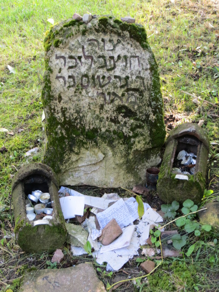 Gedenkstein für Gerschom Me’or ha-Gola auf dem Mainzer Denkmalfriedhof Das Foto zeigt einen verwitterten und bemoosten Grabstein mit hebräischen Schriftzeichen. Auf ihm liegen nach jüdischer Tradition kleine Steine zur Erinnerung an den Verstorbenen. Vor dem Stein sind Papierzettel mit Nachrichten an den Verstorbenen gestapelt.