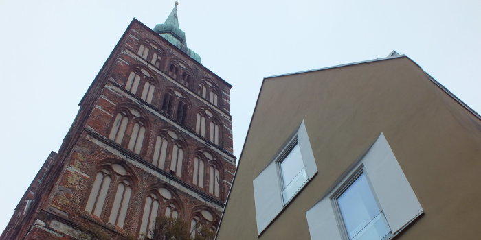 Links im Bild und im Hintergrund ragt ein gotischer Backsteinkirchturm in den Himmel. Auf der rechten Bildhälfte und eher im Vordergrund der erste Stock und das Dachgeschoss eines modernen, beige verputzten Wohnhauses