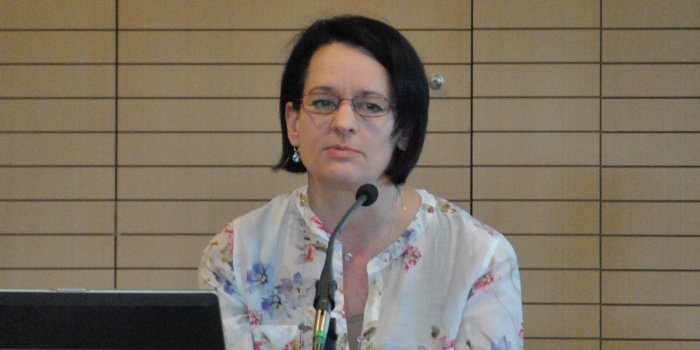 Foto: Birgit Wiedl © B. Wiedl Vor einer hölzernen Paneelwand und hinter einem Pult steht eine Frau und hält einen Vortrag. Sie trägt dunkle halblange Haare, eine schmal rot gefasste Brille und eine helle Bluse mit geblümtem Muster.