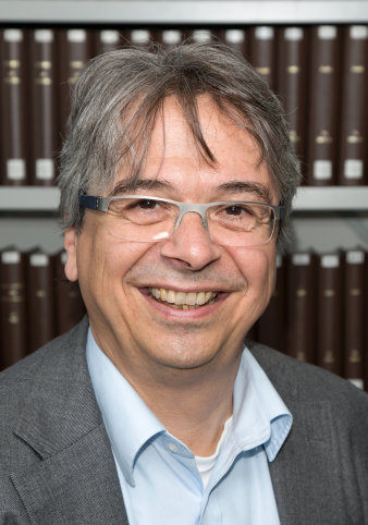 Foto: Lukas Clemens © Lukas Clemens Ein Mann mit grauer Brille und grauem Sakko steht vor einem Bücherregal und lächelt in die Kamera.