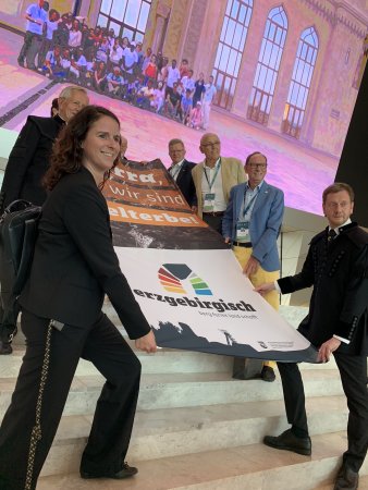 Foto: Friederike Hansell bei der Titelverleihung 2019 © IWTG Acht Personen halten auf einer Treppe stehend ein Banner, auf dem steht: "Hurra, wir sind Welterbe!"