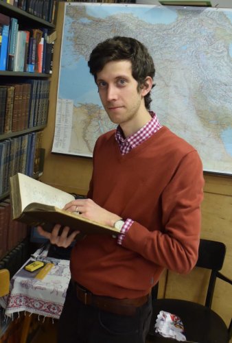 Ein Mann mit dunklen Haaren steht vor einer Landkarte. Er hält ein Buch in der Hand und blickt in die Kamera. 
