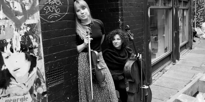 Fran & Flora Eine Frau mit Violine und eine Frau mit Cello stehen an einer Häuserwand.