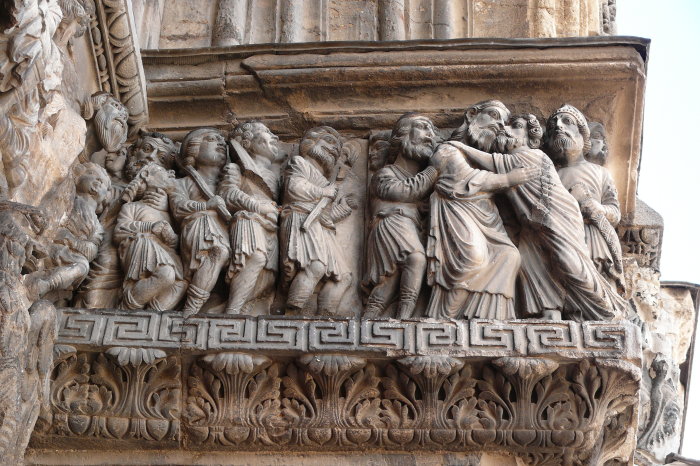 Der Judaskuss an der Abteikirche in Saint-Gilles-du-Gard (12. Jahrhundert) Die Aufnahme zeigt ein Relief an einer Kirche. Judas küsst Jesus auf die Wange, andere Jünger stehen daneben.