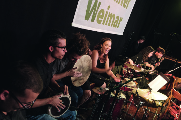 Caravan Orchestra & Choir Sechs Personen musizieren gemeinsam mit verschiedenen Gitarren und Trommeln.