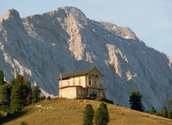 ein Haus auf einem grünen Berg vor Hochgebirgskulisse