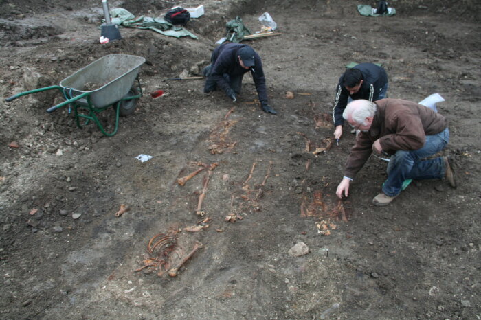 Foto einer archäologischen Ausgrabung, drei Herren kniend an einer Ausgrabungsstelle