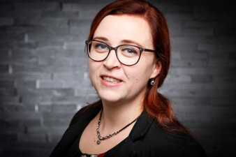 Foto: Portrait Dr. Malin Drees © Malin Drees Frau mittleren Alters mit Brille und langen, roten Haaren vor einer schwarzen Steinwand