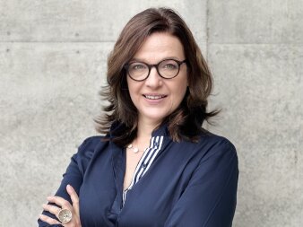 Foto: Portrait Birgit Kita © Birgit Kita Portrait einer Frau im blauen Oberteil und mit Brille vor einer hellen Steinwand
