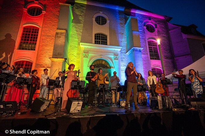 Ein Ensemble jüdischer Musiker:innen mit Instrumenten bei einem Auftritt vor einem bunt beleuchteten Gebäude