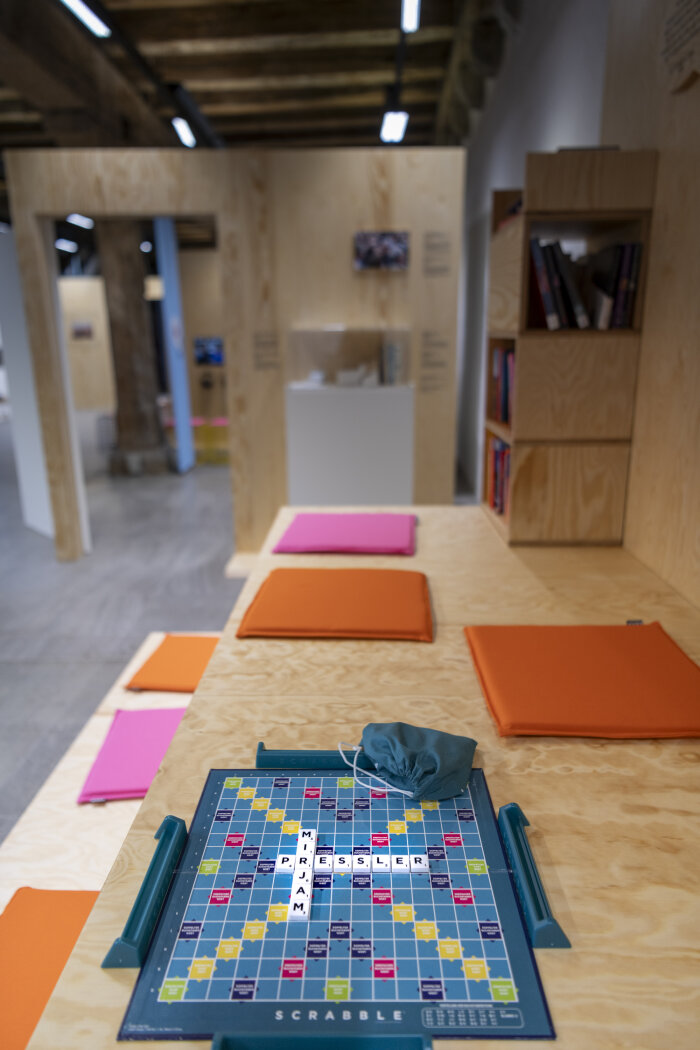 Wortspiele in der Ausstellung - Galerie Waidspeicher Ein Scrabble Spiel auf einem Holzuntergrund liegend mit bunten Kissen dabei