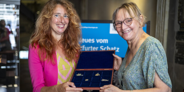 eine Frau mit langen roten Haaren und eine Frau mit kurzen blonden Haaren halten einen Schaukasten mit Schmuck