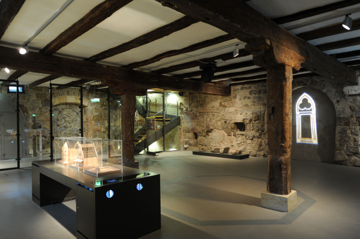 Foto: Ausstellung Alte Synagoge Erdgeschoss © Papenfuss - Atelier für Gestaltung Großer Raum mit Holzstütze in der Mitte. Verschieden Vitrinen und Ausstellungstelen im Raum verteilt.