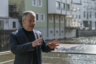 ein Mann gestikuliert mit den Händen, hinter ihm eine Ausstellungstafel und Blick aufs Wasser und eine Häuserzeile