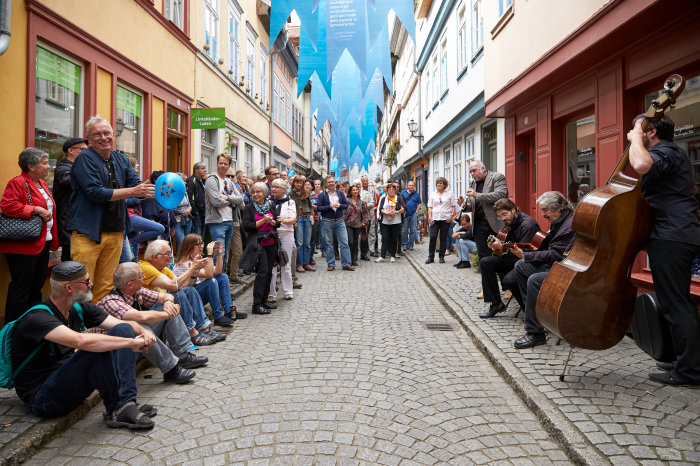 Viele Menschen sitzen und stehen auf der Krämerbrücke, sie schauen einer Band dabei zu, wie sie musizieren. 