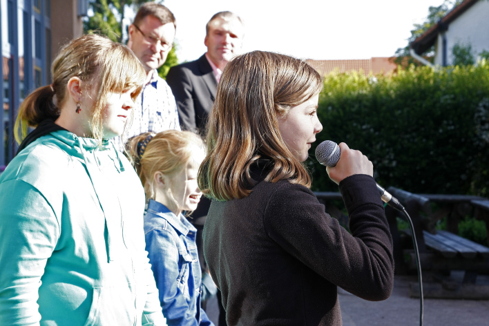 Mädchen singt in ein Mikrofon, im Hintergrund der Rest des Chores und Zuhörer.