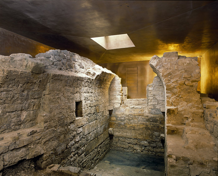 Mauerreste der mittelalterlichen, jüdischen Mikwe. Reste eines kleinen Raumes mit Tonnengewölbe, umgeben von Ausschnitten des bronzefarbenen Schutzbaus.