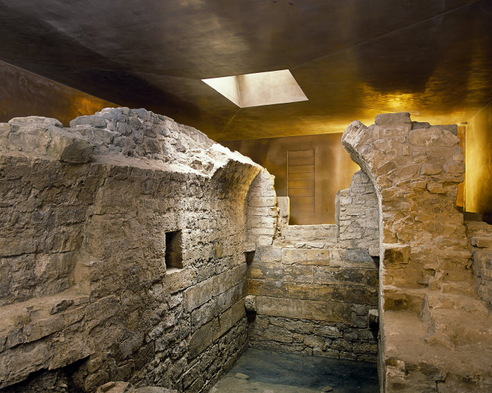 Foto: Mikwe Erfurt © Peter Seidel Mauerreste der mittelalterlichen, jüdischen Mikwe. Reste eines kleinen Raumes mit Tonnengewölbe, umgeben von Ausschnitten des bronzefarbenen Schutzbaus.