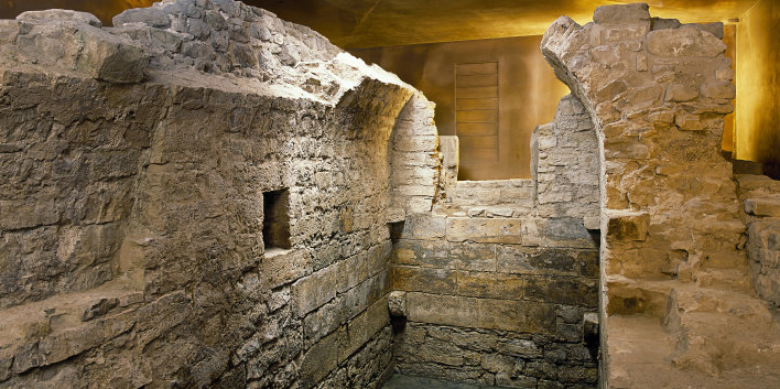 Foto: Mikwe Erfurt © Peter Seidel Mauerreste der mittelalterlichen, jüdischen Mikwe. Reste eines kleinen Raumes mit Tonnengewölbe, umgeben von Ausschnitten des bronzefarbenen Schutzbaus.