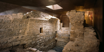 Mauerreste der mittelalterlichen, jüdischen Mikwe. Reste eines kleinen Raumes mit Tonnengewölbe, umgeben von Ausschnitten des bronzefarbenen Schutzbaus.