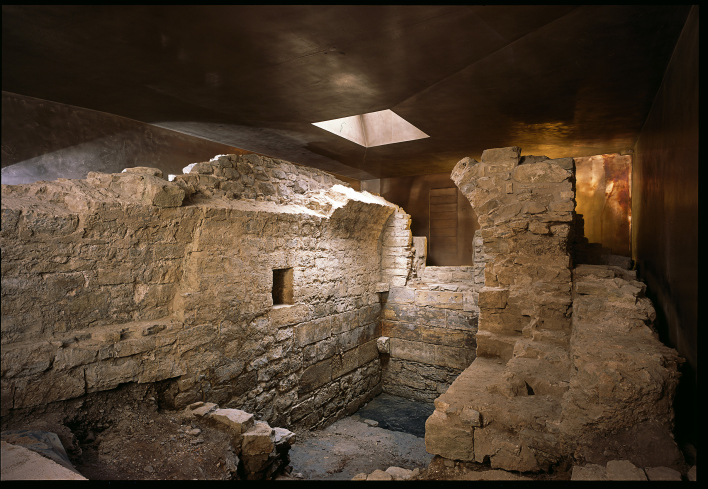 Mauerreste der mittelalterlichen, jüdischen Mikwe. Reste eines kleinen Raumes mit Tonnengewölbe, umgeben von Ausschnitten des bronzefarbenen Schutzbaus.