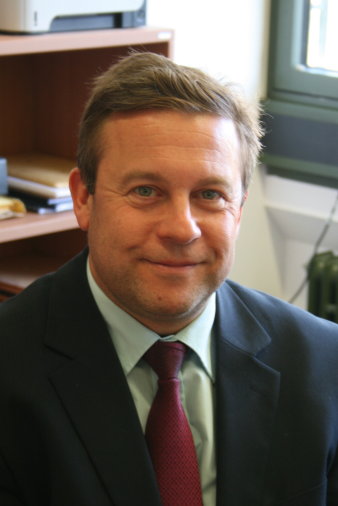 Ein Mann mit kurzen braunen Haaren lächelt freundlich in die Kamera. Er trägt einen grauen Anzug, ein weißes Hemnd und eine rote Krawatte. Im Hintergrund ist ein Büro zu sehen.