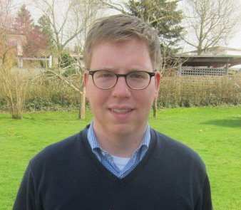 Foto: Johann Deil © Johann Deil Ein junger Mann mit schwarzer Brille und kurzen braunen Haaren steht in einem sonnenbeschienenen Garten und blickt in die Kamera. Er trägt einen dunkelblauen Pullover und ein blauweiß kariertes Hemd.