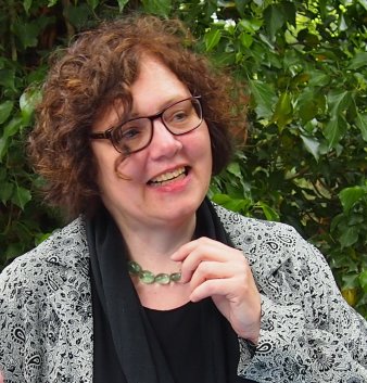 Eine Frau mit kurzen, gelockten braunen Haaren und Brille lacht und redet mit jemandem rechts vom Fotgrafen. Im Hintergrund sind grüne Blätter zu sehen.