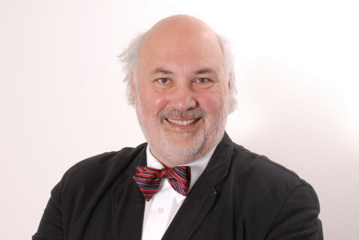 Wolfgang Schneider Ein Herr mit kurzem grauen Vollbart und Haarkranz sitzt vor einem weißen Hintergrund. Er trägt zum dunkelgrauen Jackett und weißen Hemd eine rot-blau-weiß gestreifte Fliege und lächelt in die Kamera.