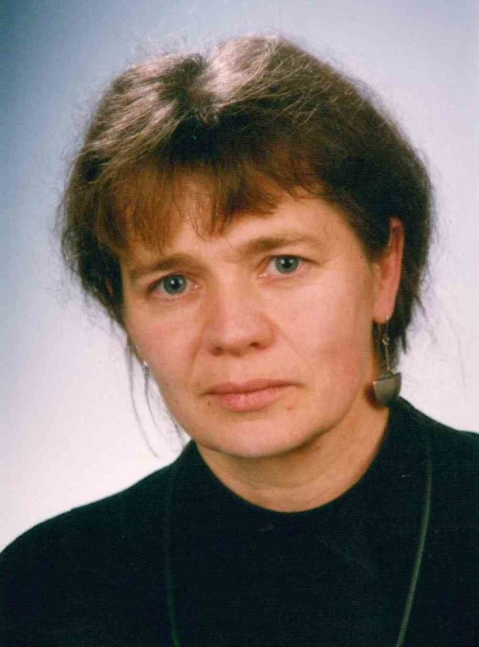 Angelika Schneider Zu sehen ist der Kopf und Oberkörper einer Frau vor neutralem Hintergrund. Sie blickt in die Kamera, hat dunkles hochgestecktes Haar mit einem Pony, im linken Ohr trägt sie einen langen metallenen Ohrring.