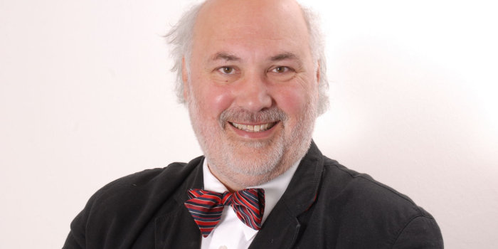 Foto: Wolfgang Schneider © Universität Hildesheim Ein Herr mit kurzem grauen Vollbart und Haarkranz sitzt vor einem weißen Hintergrund. Er trägt zum dunkelgrauen Jackett und weißen Hemd eine rot-blau-weiß gestreifte Fliege und lächelt in die Kamera.