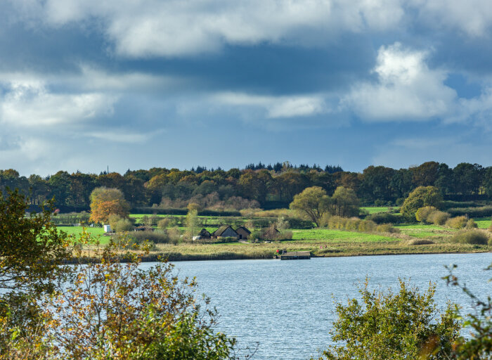 Blick auf den Siedlungsbereich von Haithabu mit rekonstruierten Häusern und Landebrücke in der Bildmitte und dem umgebenden, Baum bestandenen Wall im Hintergrund Ein Fluss, dahinter eine Wiese mit Hütten und Wald