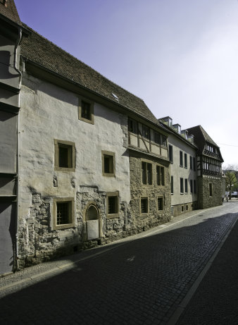 Eine mit Kopfstein gepflasterte Straße im Sonnenlicht. Eine Reihe unterschiedlich alter Häuser ist zu sehen, eines mit Fachwerk, eines aus grob verputztem Mauerwerk, ein zeitgenössisches Gebäude.