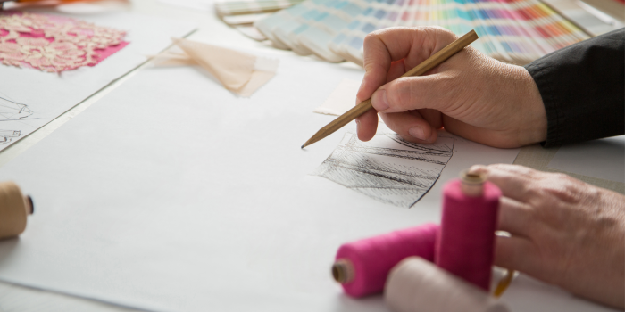 Foto: Im Workshop werden eigene Modekreationen entworfen und umgesetzt. © E. Fuckel Eine Hand mit Bleistift zeichnet einen Rock. Im Hintergrund Farbfächer und Nähgarn.