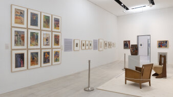 Ein Ausstellungsraum mit Gemälden an den Wänden rechts zwei Stühle aus Holz