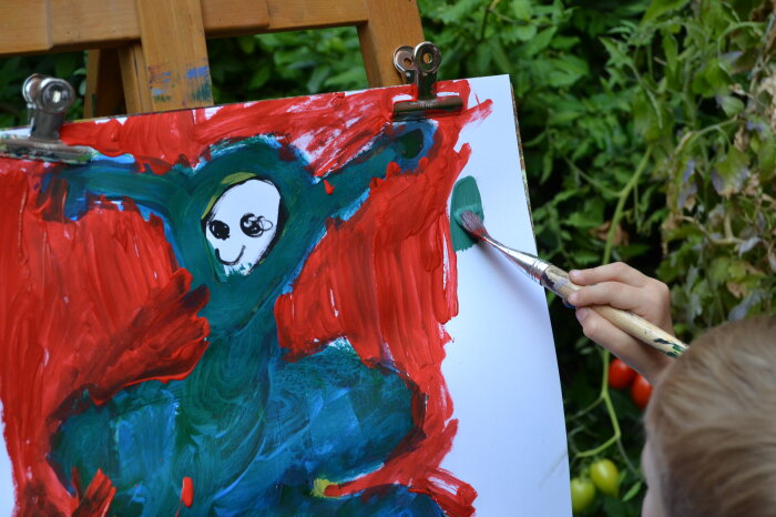 Die Teilnehmenden werden gemeinsam kreativ. Eine Leinwand mit einer blau-roten Kinderzeichnung, rechts eine Hand die einen Pinsel hält