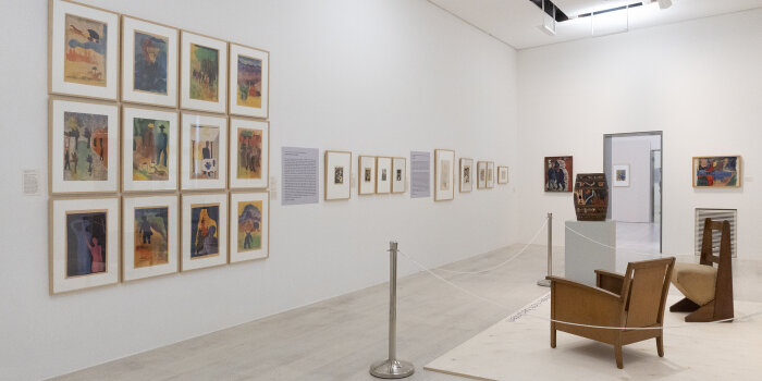 Ein Ausstellungsraum mit Gemälden an den Wänden rechts zwei Stühle aus Holz