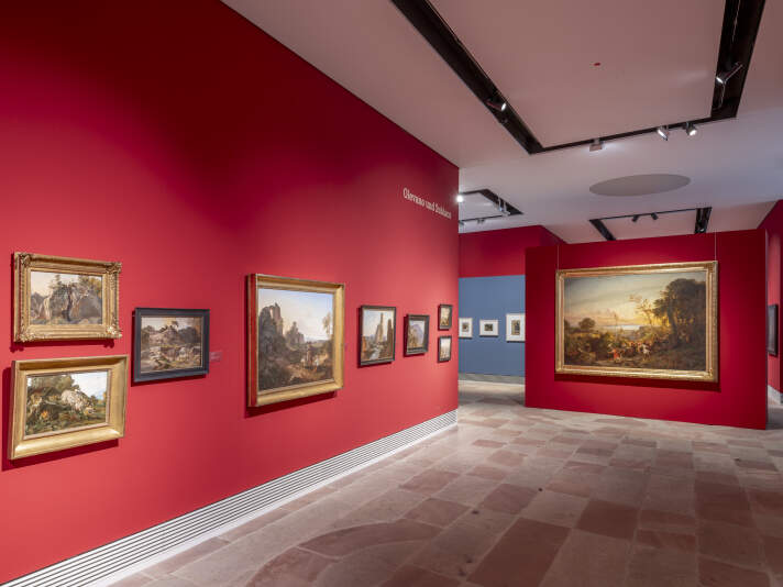 Ein Ausstellungsraum mit roten und blauen Wänden an den Gemälde hängen.